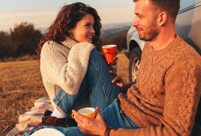 Beautiful young couple dating in their thirties and enjoying picnic time on the sunset. They drinking tea and sitting in a meadow leaning against a old fashioned car.