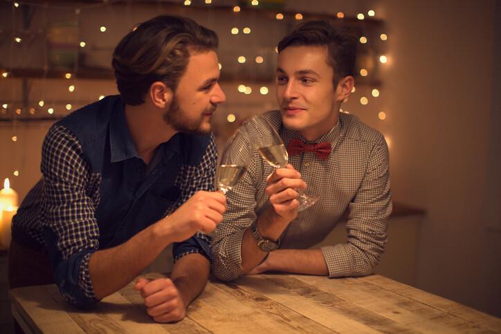 Loving gay couple having romantic date, drinking champagne. Domestic kitchen. Handsome caucasian man looking at each other. Evening scene lit by candle and string lights.