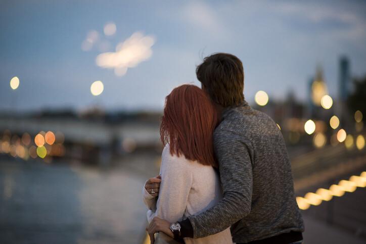 A couple is seen from behind while hugging and watching fireworks at sunset with city lights in the background.