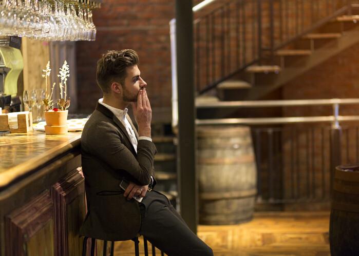 A young man is dressed smarty at a bar waiting, he looks fed up as if he has been stood up on a date. One hand is on his face and the other is holding his smart phone.