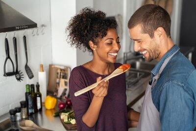 Young couple tasting tomato sauce while cooking in the kitchen. Cheerful man and smiling woman holding spatula in hand ready to taste red sauce. Multiethnic couple cooking together at home. No arguments.