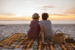 Lesbian couple with backs to camera, sitting on a blanket on the beach, looking towards the ocean